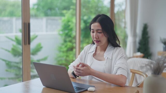 Focused young Asian woman gesturing with hands while talking on a video call using a laptop, working remotely at a wooden table in a bright modern home with a garden view.