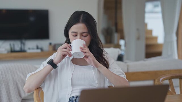 Happy young Asian woman drinking hot coffee from a white mug while taking a break from her laptop in a modern living room at home.