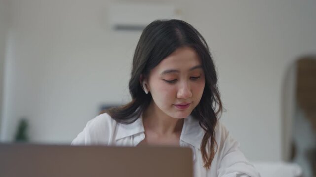 Close up portrait of Asian woman with wireless earbuds focusing on laptop screen, working and searching for information online at home in a bright minimal roomlaptop, earbuds, working, close up, home,