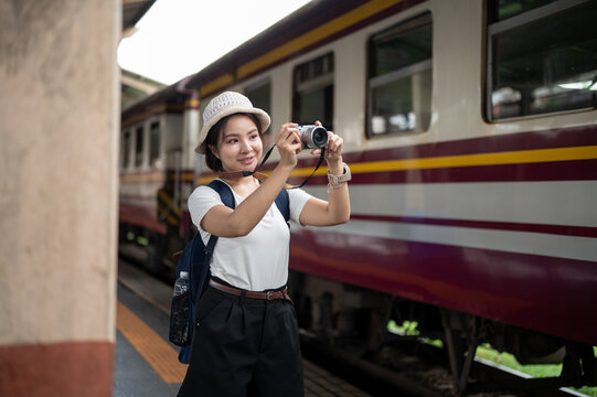 Asian woman traveler taking photos with camera beside a passenger train at railway station platform.