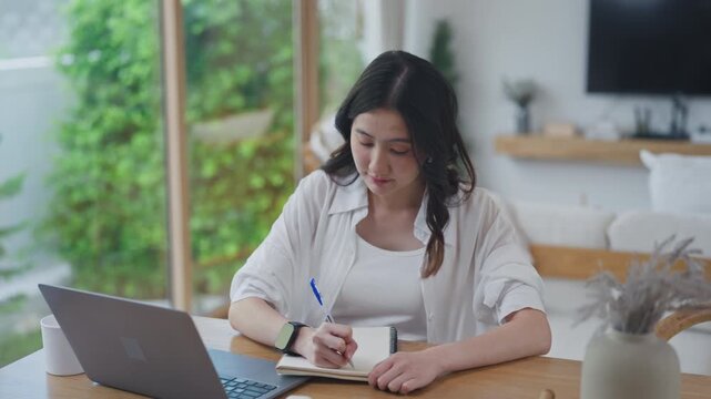 Asian woman taking notes by hand and planning her finances at home, writing in notebook beside laptop, organizing tasks and setting goals in a bright minimal room