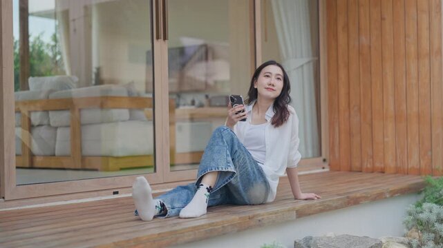 Asian woman sitting on wooden terrace holding smartphone, taking a break and relaxing at modern minimal home, enjoying slow living and a calm weekend moment