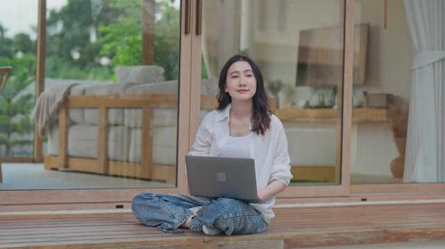 Asian woman sitting cross legged on wooden terrace using laptop, working from home in a calm minimal space, balancing work and relaxation in a slow living lifestyle