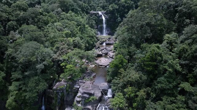 A drone flying over a waterfall in a deep green forest provides an aerial, top-down view of a National Park in Laos PDR.