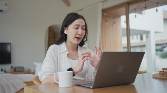 Asian woman wearing earbuds typing on laptop and using AI assistant for online learning at home, talking and working with chatbot technology in a bright modern room