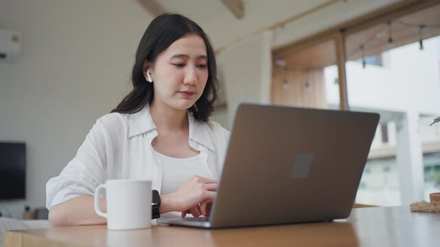 Asian woman wearing earbuds on online video call, working from home on laptop with AI assistant support, talking and typing during a remote meeting in a modern room