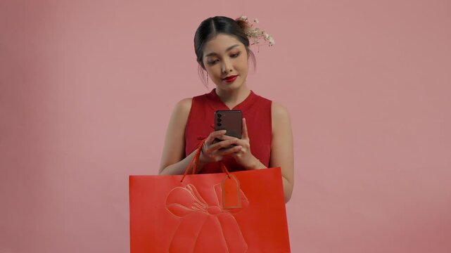 Shocked young Asian woman in a traditional cheongsam dress is holding smartphone and shopping bags, isolated by pink background.
