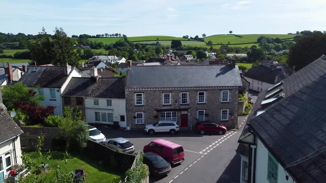 English village houses and parked cars line a narrow Colyton street with green East Devon hills beyond, viewed from a slowly rising drone.