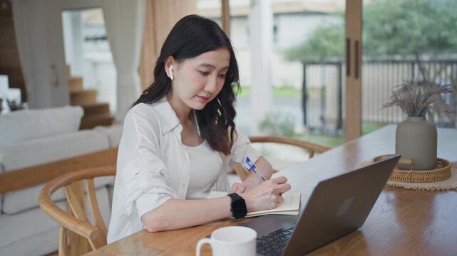 Asian woman wearing earbuds typing on laptop with deep focus, working from home and concentrating on her tasks at a wooden table in a bright modern room