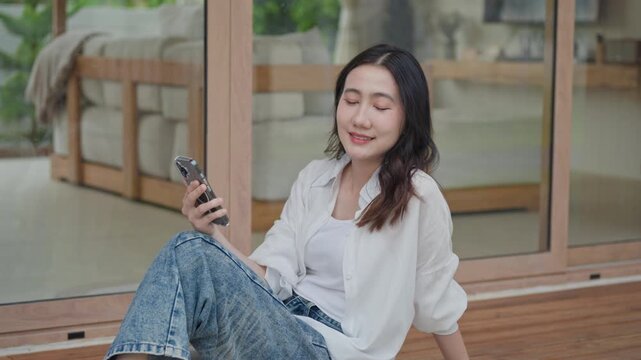 Asian woman sitting on wooden floor holding smartphone, taking a calm break at modern home, enjoying a peaceful mindful moment of slow living and self care