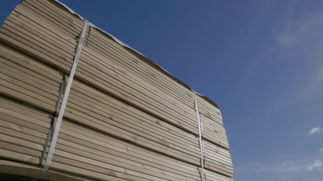 Large bundles of cut wooden boards are stacked on pallets outdoors under a blue sky, ready for storage, shipping, or construction supply