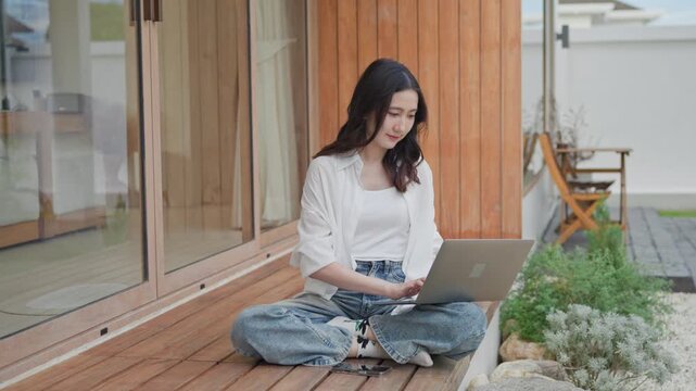 Asian woman sitting cross legged on wooden terrace working on laptop outdoors beside a home garden, enjoying flexible remote work in a calm outdoor space at modern home