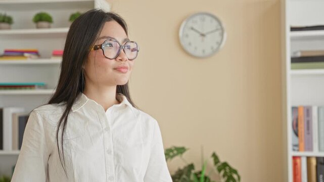 Young woman wearing round glasses gazes to the right, white shirt visible, standing by bookshelf, potted plant and wall clock on beige wall in a building; calm focus.