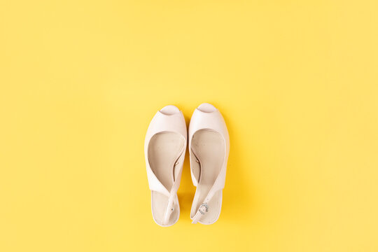A pair of light-colored, open-toe heeled shoes are centered on a solid yellow background, showcasing a minimalist and stylish footwear option.
