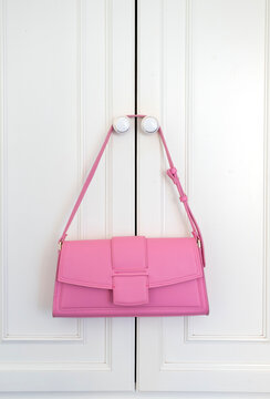 A vibrant pink rectangular shoulder bag with a magnetic flap closure hanging from white closet knobs. The clean, bright lighting emphasizes its modern silhouette and bold, feminine pop of color.
