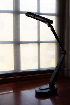 A sleek black LED desk lamp sits on a polished wooden surface against a backlit window with soft diffused light. The minimalist silhouette and articulated arm create a modern, professional aesthetic.