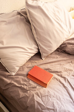 A cozy, high-angle shot of a small red vintage book resting on unmade purple-grey bedding. The soft natural morning light creates a peaceful, inviting atmosphere perfect for a slow lifestyle theme.