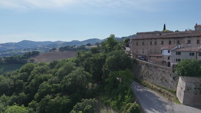 Borgo collinare con case in pietra e tetti in terracotta, dominato da una torre campanaria e circondato da alberi e campagna verde sotto cielo azzurro
