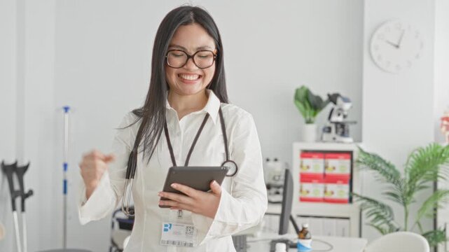 Woman doctor in white coat smiling and tapping a tablet, fist pump gesture with stethoscope visible in a medical office building; confidence success.