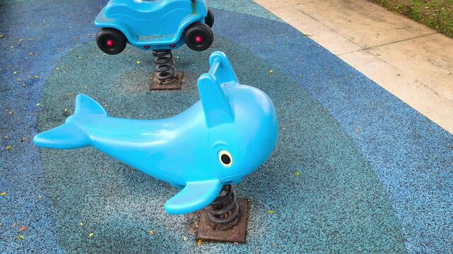 A static shot capturing an empty blue dolphin-shaped spring rider swaying slightly on a colorful EPDM safety rubber floor in an outdoor children's playground park. Video transition, 4k, 60 fps