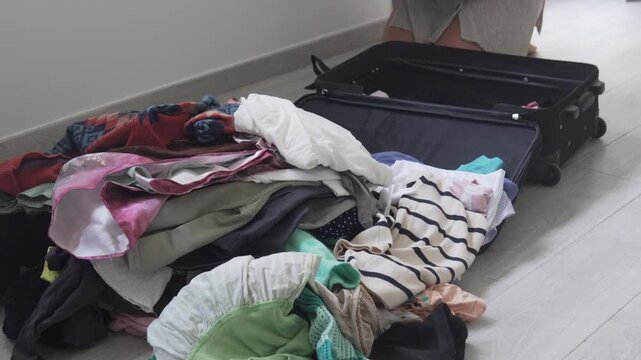 Open suitcase and pile of clothes on the floor during travel packing at home, vacation preparation and relocation concept