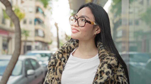 Woman wearing tortoiseshell glasses and leopard coat smiling and looking up on an urban street near parked cars and glass storefronts; optimism.