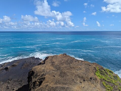 Panoramic view of a tropical ocean from a Halona Blowhole Lookout. Rugged volcanic lava rock in the foreground, turquoise and deep blue water with white sea foam, light blue sky with cumulus clouds.