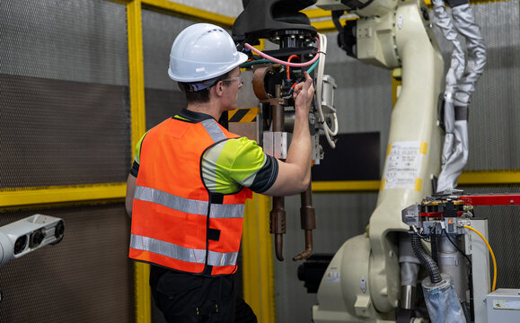 Industrial Robot Technician: A male technician in a hard hat and safety vest supervises an industrial robotic arm in a factory setting, emphasizing precision engineering and automation.