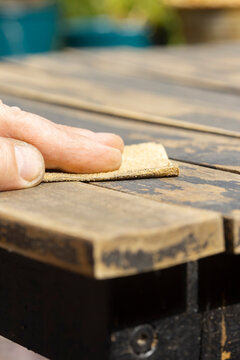 Man sanding loose peeling black paint off a wooden outdoor patio table. Garden DIY chore concept