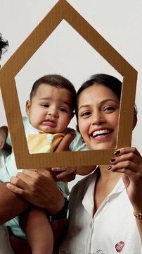 Indian young parents and baby boy looking through house shaped cardboard cutout to promote real estate investment, isolated on white background, representing happy Asian family and housing concept