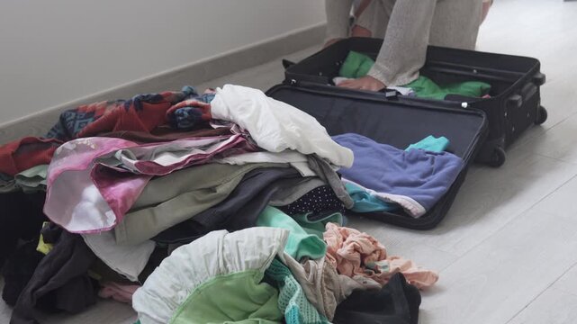 Woman packing an open suitcase with a large pile of clothes on the floor at home. Travel preparation, vacation packing, relocation, moving house, wardrobe organization and messy luggage concept