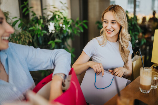 Two young women smiling as they look through shopping bags at a cafe. A cheerful shopping moment between friends sharing purchases over coffee.