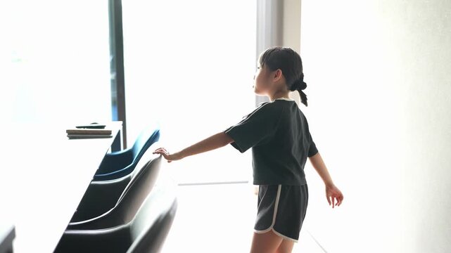 Little girl walks toward bright window with curiosity. Her dark hair ties neatly behind. She wears simple black outfit, shorts. Sunlight floods room, soft and warm. Gentle movement, quiet, peaceful