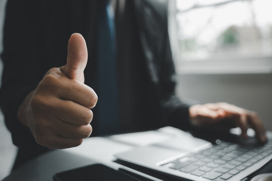 A closeup of a man hand pose with a success sign gesture, the businessman gives a thumbs up sign for approval while working on a laptop at his modern office desk