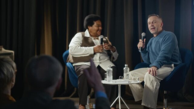 Senior Caucasian man raising hand to ask question during panel discussion with Black woman and Caucasian man on stage. Ideal for corporate communication and educational event videos, handheld
