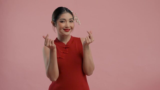 Cute young Asian woman in a traditional cheongsam qipao dress doing love heart finger gesture, isolated by pink background.