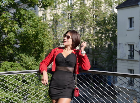 Young Asian woman wearing a black mini dress, red cardigan, and black leather boots leans against a metal railing on bridge with a building in the background