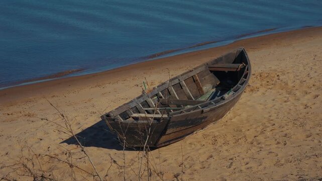 Wooden rowboat rests on golden sand. Calm blue waters lap the shore. Sunlight glints on the boat's weathered wood. Dried grasses grow near the vessel. This peaceful scene evokes solitude by the sea