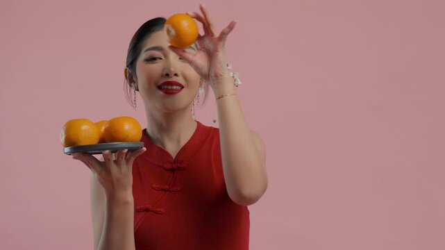 Happy young Asian woman in a traditional cheongsam dress posing while holding mandarin oranges on isolated pink background.