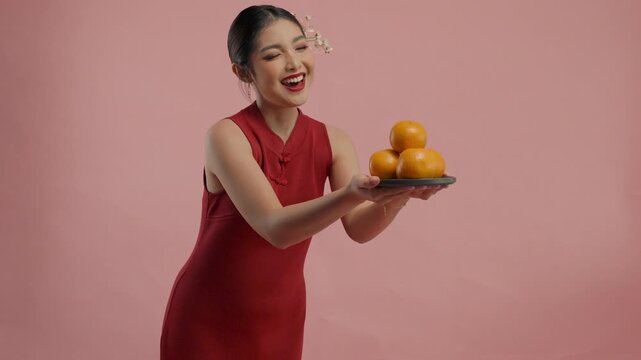 Smiling young Asian woman in a traditional cheongsam dress is giving mandarin oranges, isolated by pink background.