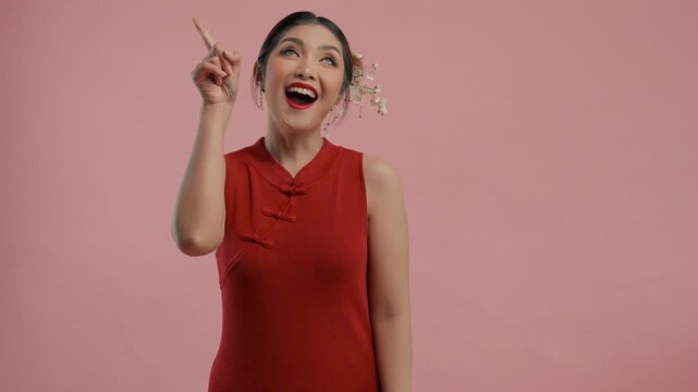 Beautiful young Asian woman in a traditional cheongsam qipao dress pointing to the copy space upwards, isolated by pink background.