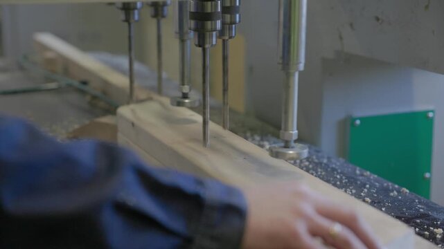 Close-up of a multi-spindle woodworking drill boring precise holes into a timber plank in a workshop, with flying sawdust and shallow depth of field