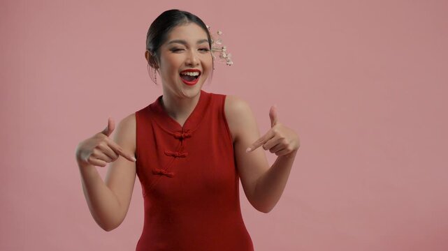 Pretty young Asian woman in a traditional cheongsam qipao dress pointing to the copy space below, isolated by pink background.