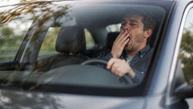 Tired male driver yawning while driving car. Man struggling with sleepiness on the road, fatigued and needing rest