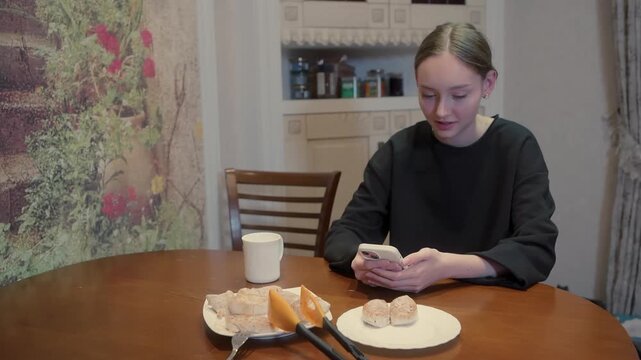 Girl sitting at wooden table reading book with coffee mug, cheese, bread, enjoying casual breakfast in cozy kitchen interior with floral wallpaper.