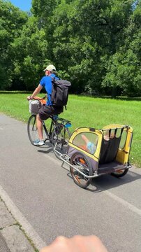 Father riding a cargo bike on a park pathway with greenery and trees in the background, calm and leisurely third-person perspective from behind.