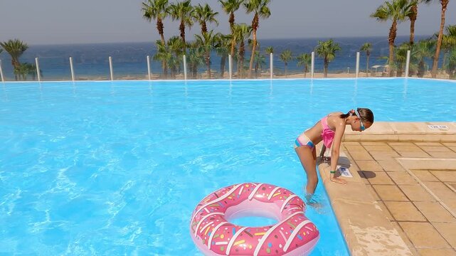 Young girl stands on pool edge ready to dive into bright blue water with inflatable pink donut float nearby, palm trees, ocean, sunny resort setting.
