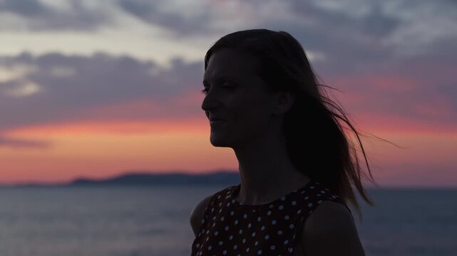 Silhouette profile of a smiling young woman in a polka dot dress against a colorful sunset sky by the sea