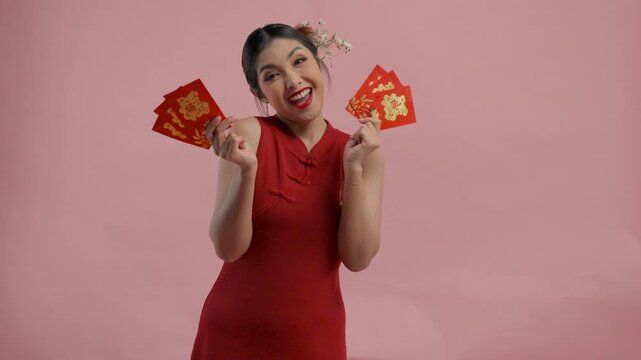 Cheerful young Asian woman is smiling and holding angpao or red packet monetary gift, isolated by pink background.