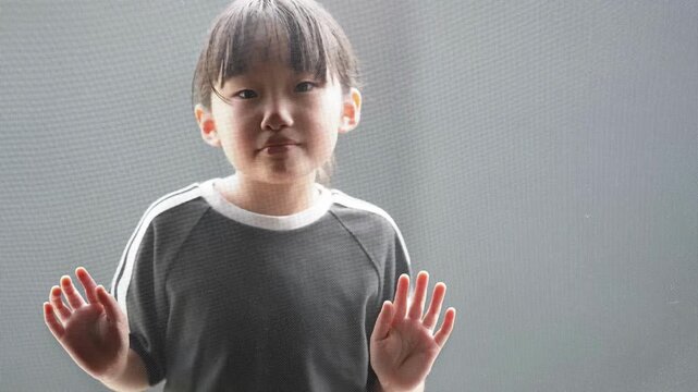 Little girl stands near window screen. Hands raised, eyes calm, expression neutral. Soft light illuminates her quiet pose. Ideal for family, children, or quiet moments concept